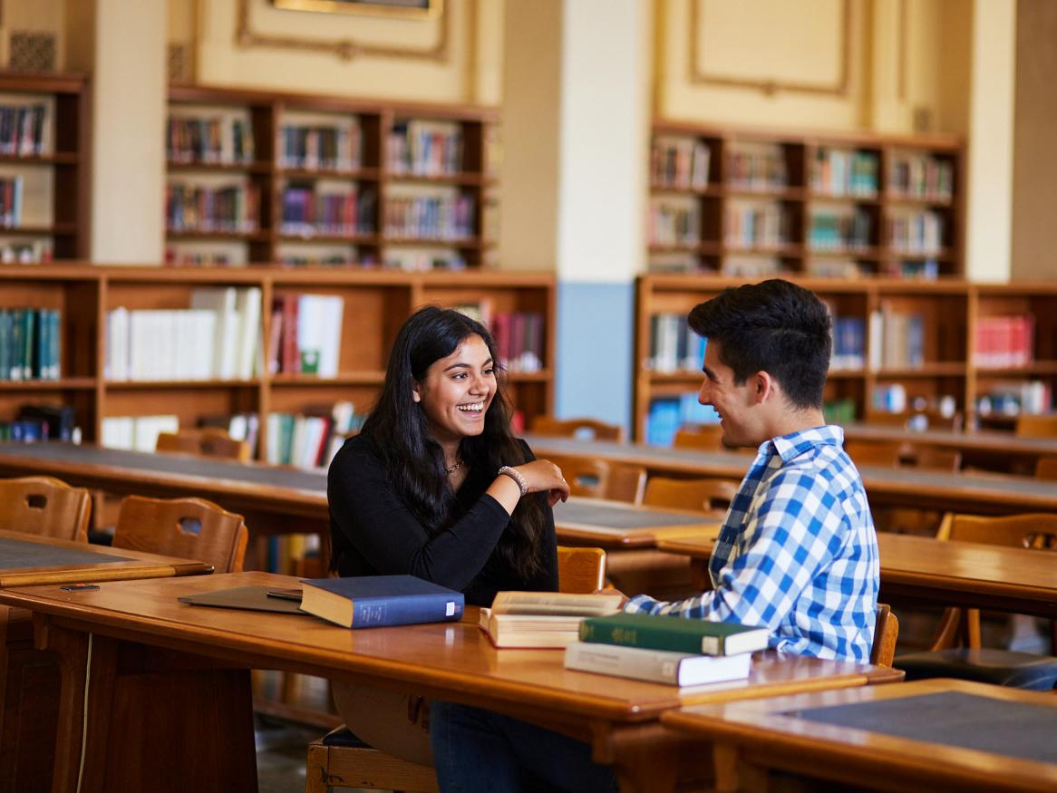 Two students sitting in the Reading Room, chatting and smiling at each other
