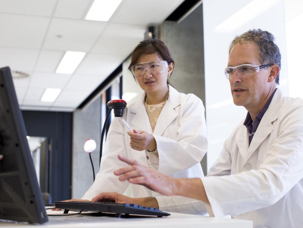 Two researchers wearing labcoats in front of a computer, gesturing
