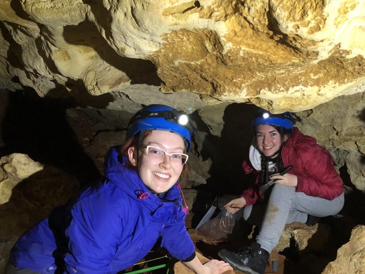 Liz Reed image - two female presenting students inside a cave with some archeological equipment, smiling at the camera