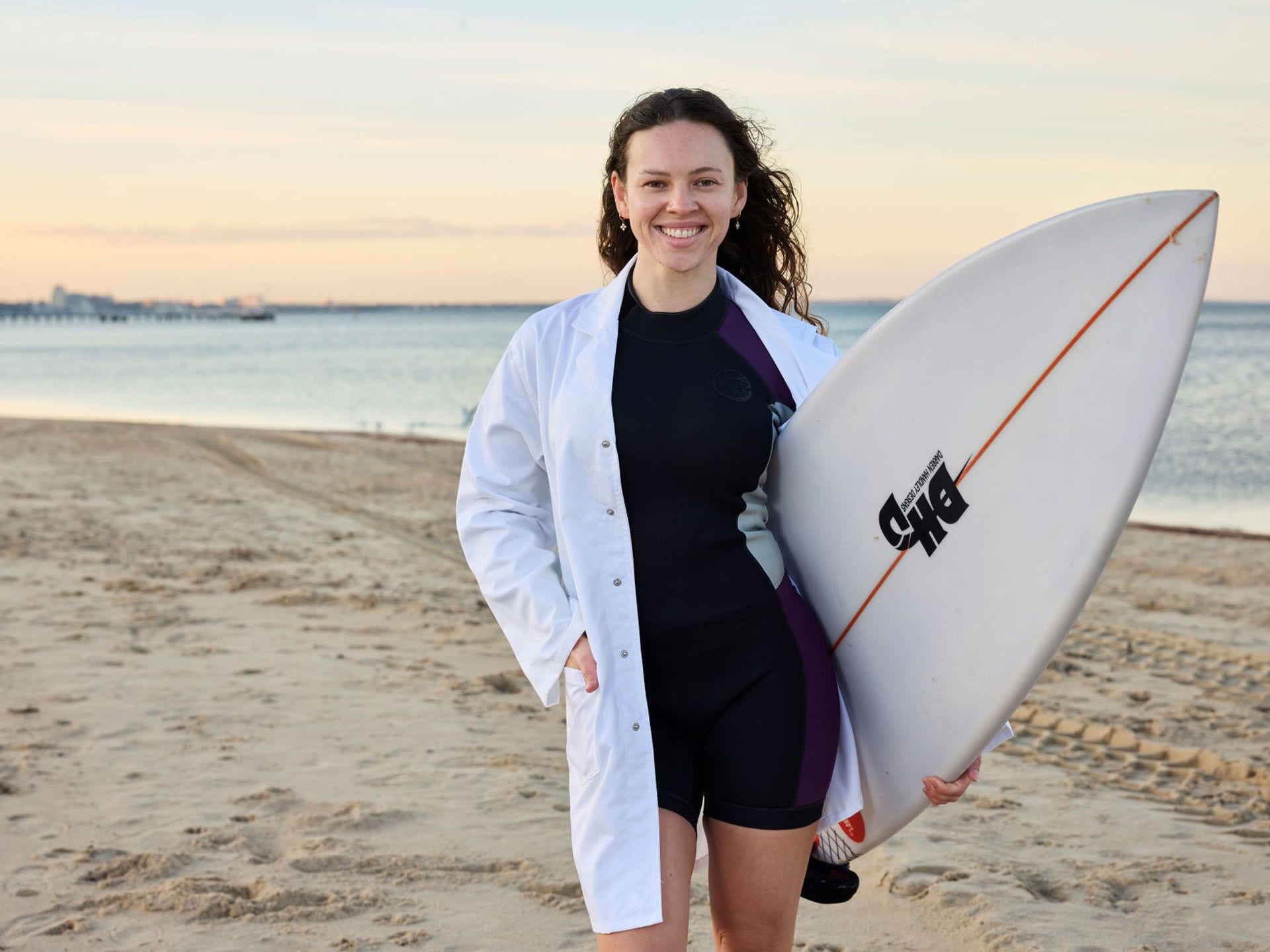 Woman on a beach holding a surfboard wearing a wetsuit with a white lab coat over it