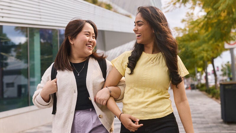 Two university students walking arm-in-arm outdoors, laughing