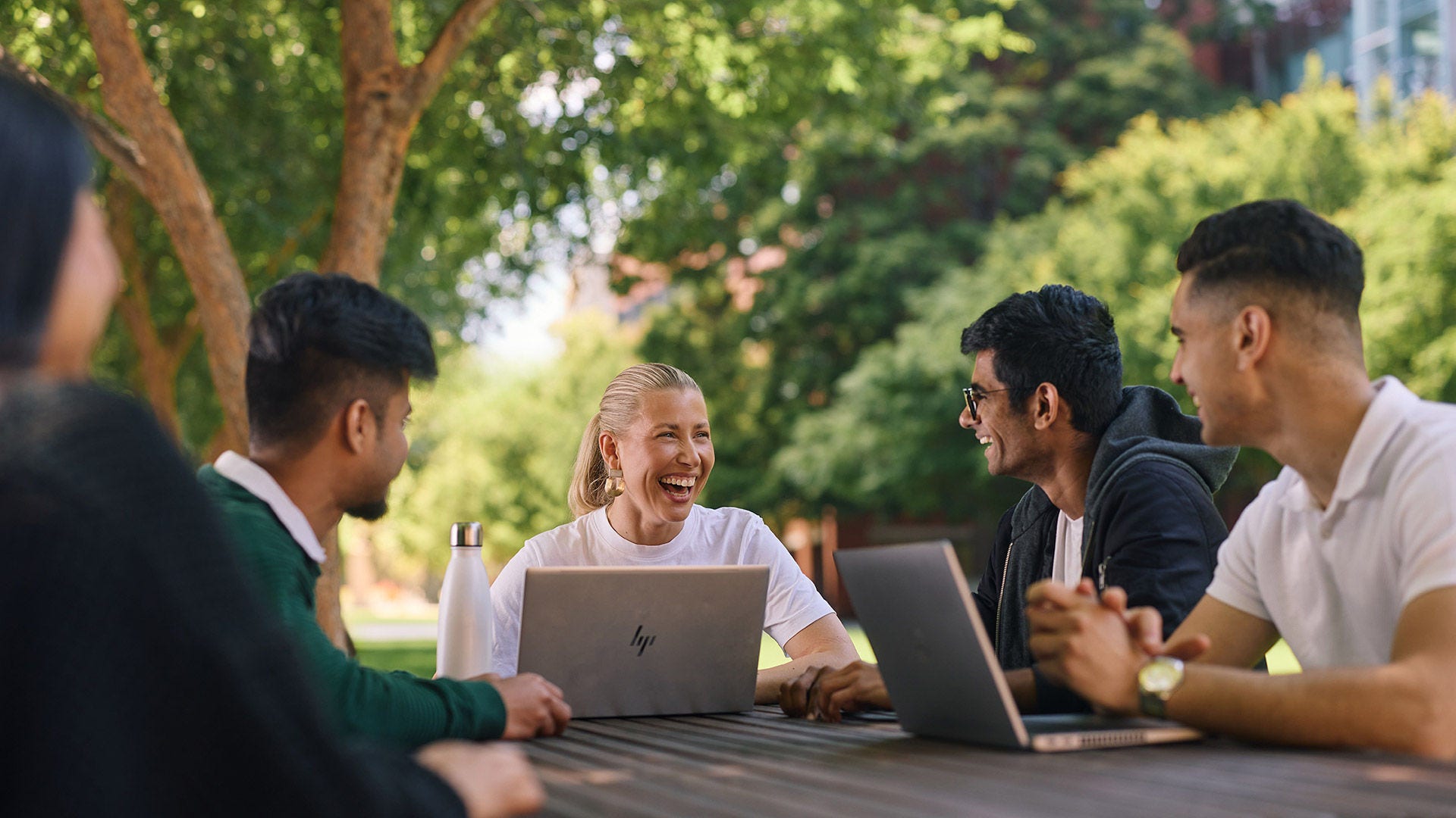 A group of students laughing together at an outdoor table with laptops, surrounded by trees.