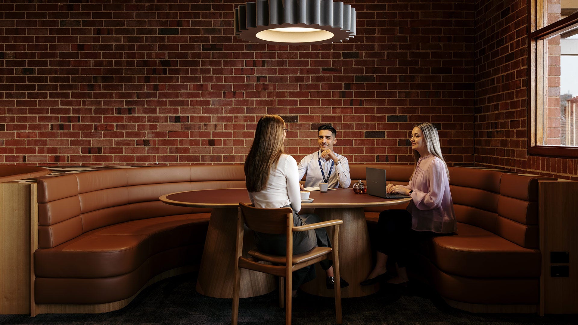 Three people meeting at a round table in a modern lounge with brick walls and curved leather seating