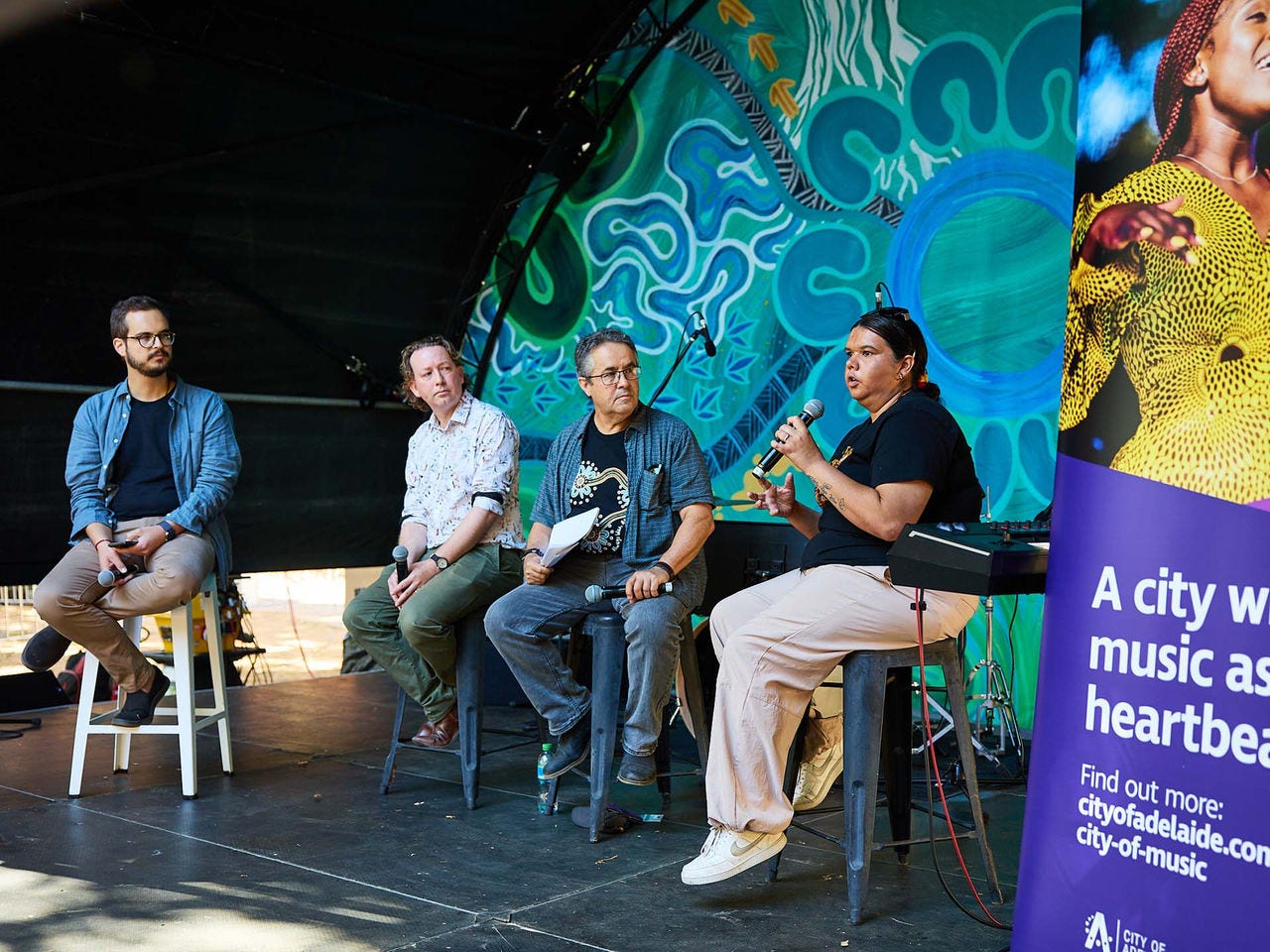 Four panellists on stage during a music discussion, with an Indigenous artwork backdrop and a City of Adelaide banner visible.