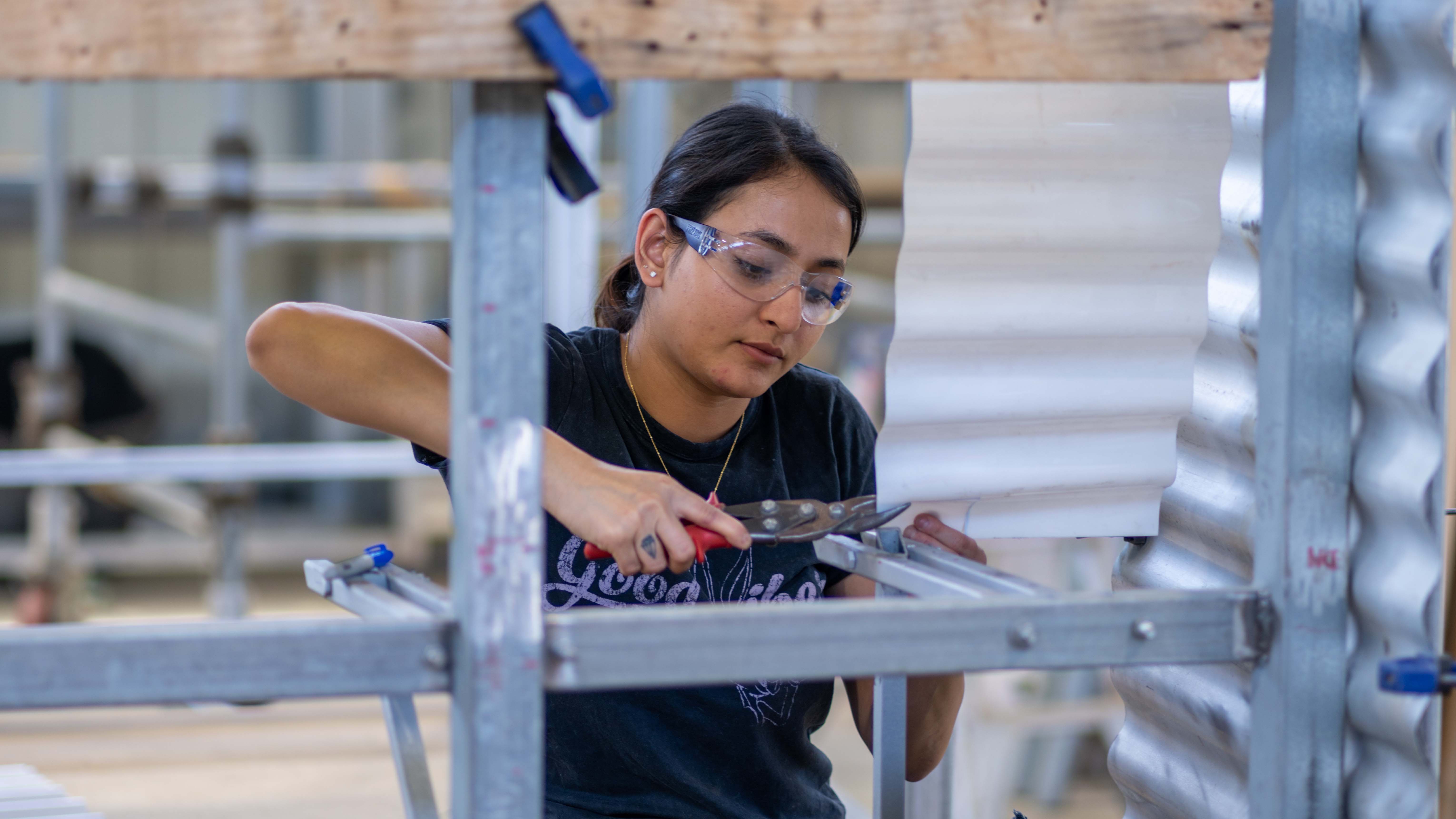 A young woman wearing safety glasses and a black T-shirt uses red-handled tin snips to cut corrugated metal sheeting on a workshop scaffold, with more sheet metal panels visible in the background