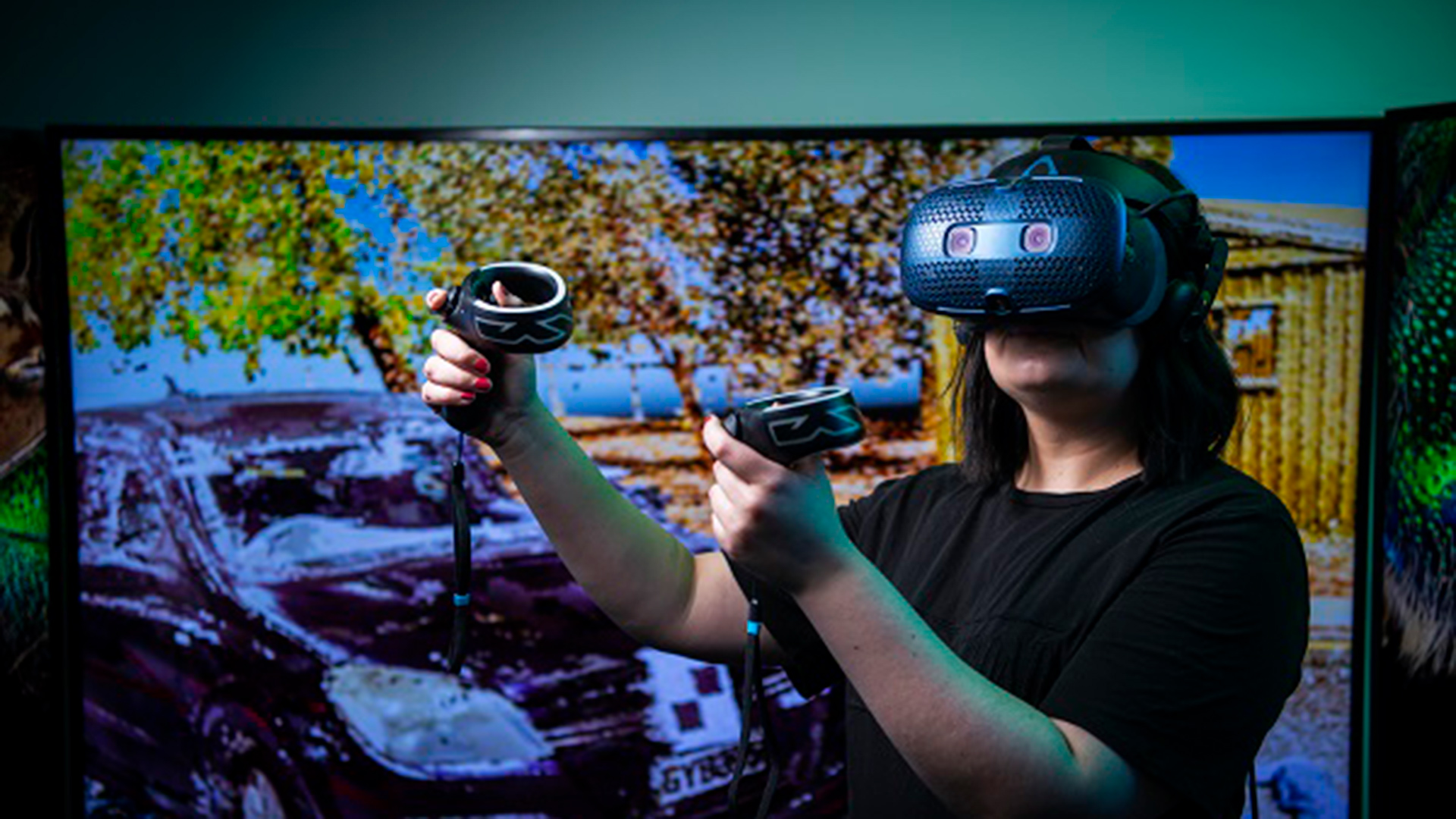 Person using VR goggles and hand held devices with a screen in the background