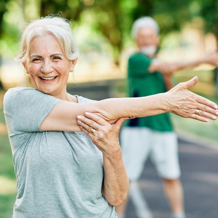 Stock image of an older woman in a park stretching arm across chest and smiling.