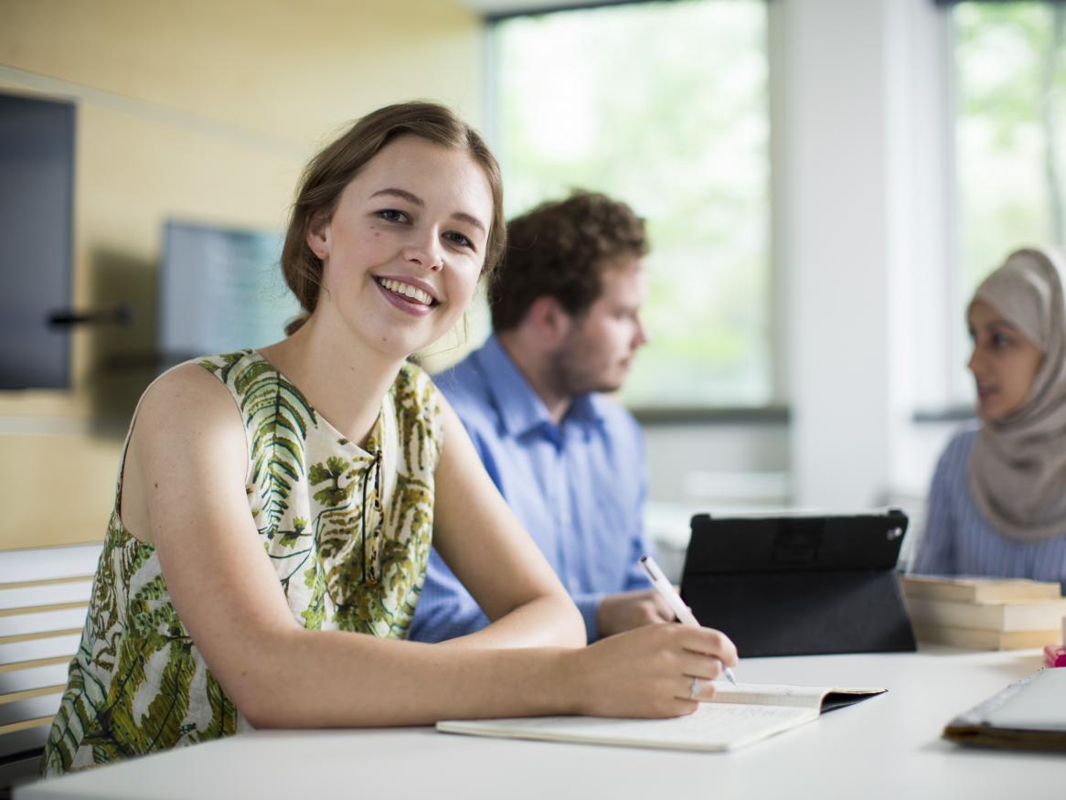 Image of a female student writing, facing the camera and smiling. Two other students are sitting nearby.