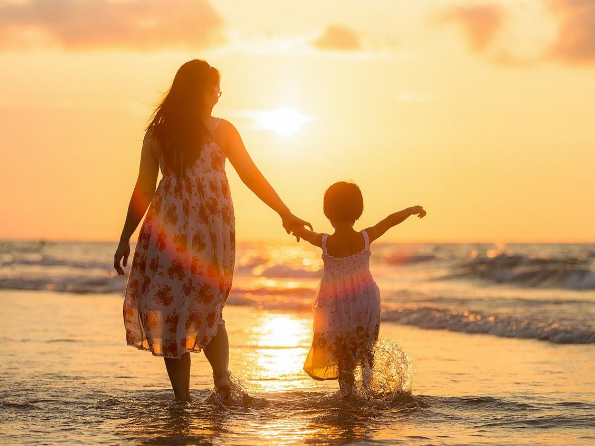A mum holds their child hand on the beach, with the sun setting.