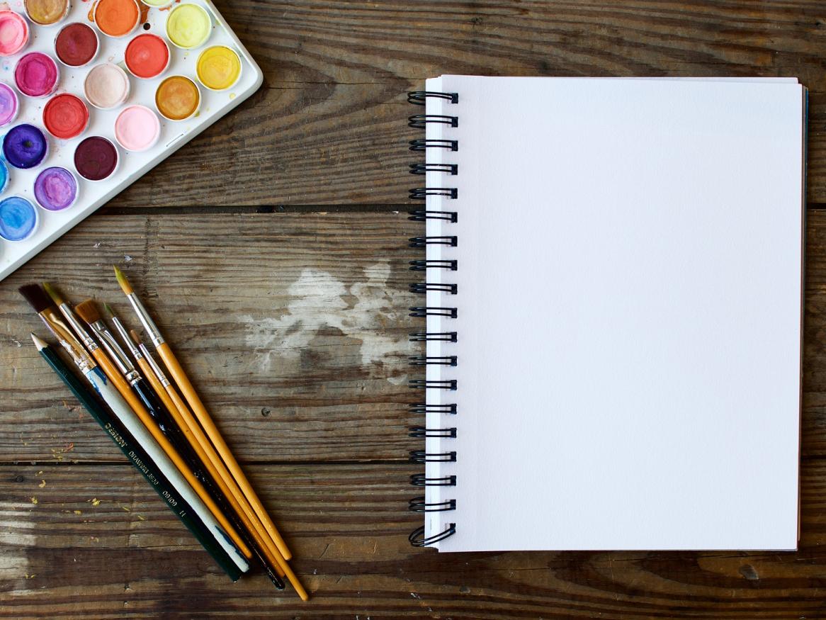 A set of watercolours, paintbrushes and a notebook on a wooden desk.