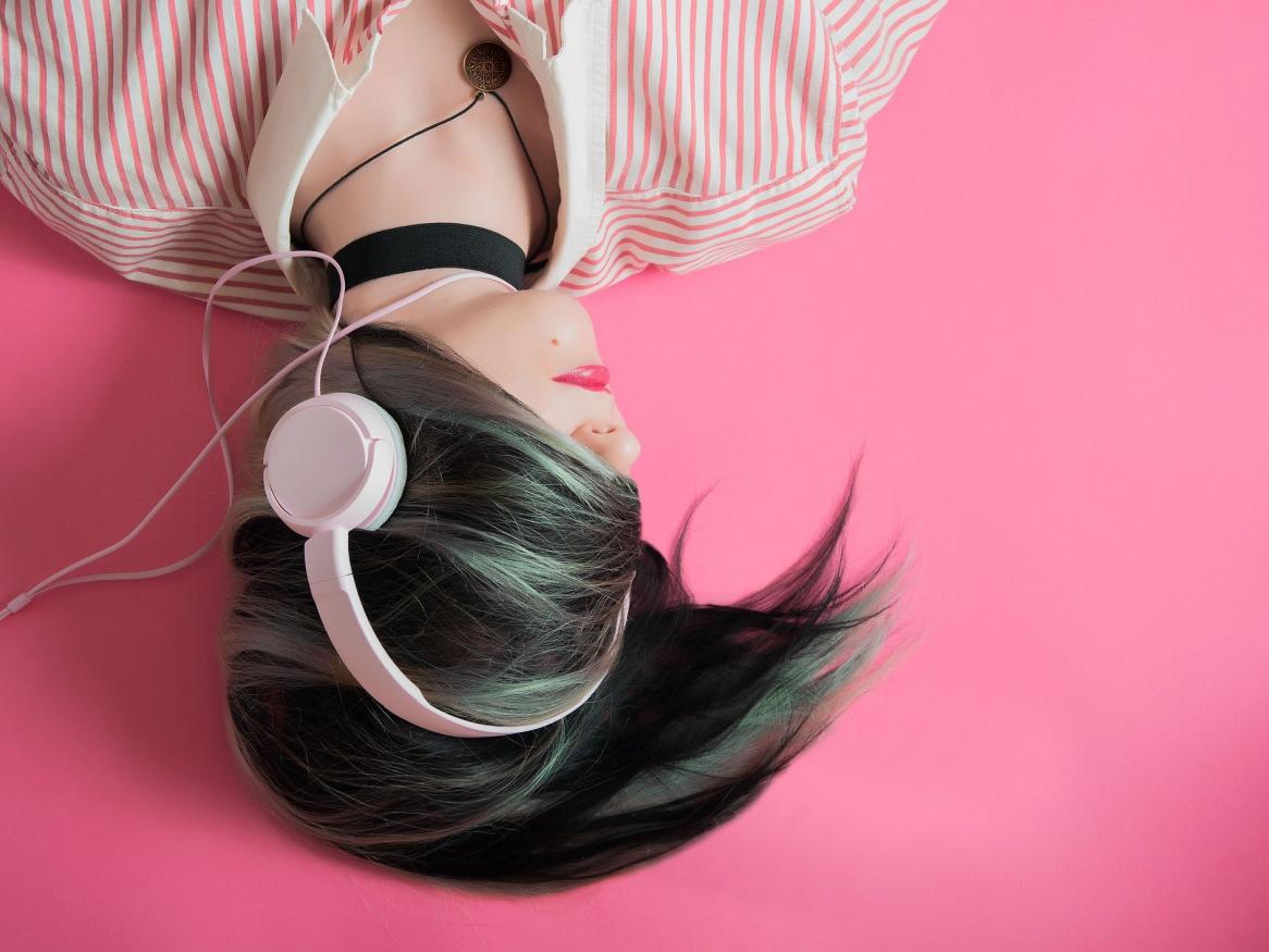 A young woman with dark hair wearing light pink headphones and a striped white and pink shirt on a deep pink background