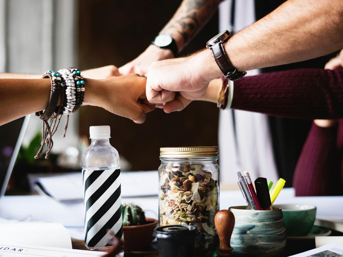 A group of people fistbump over some work, a bottle and a jar on a table.