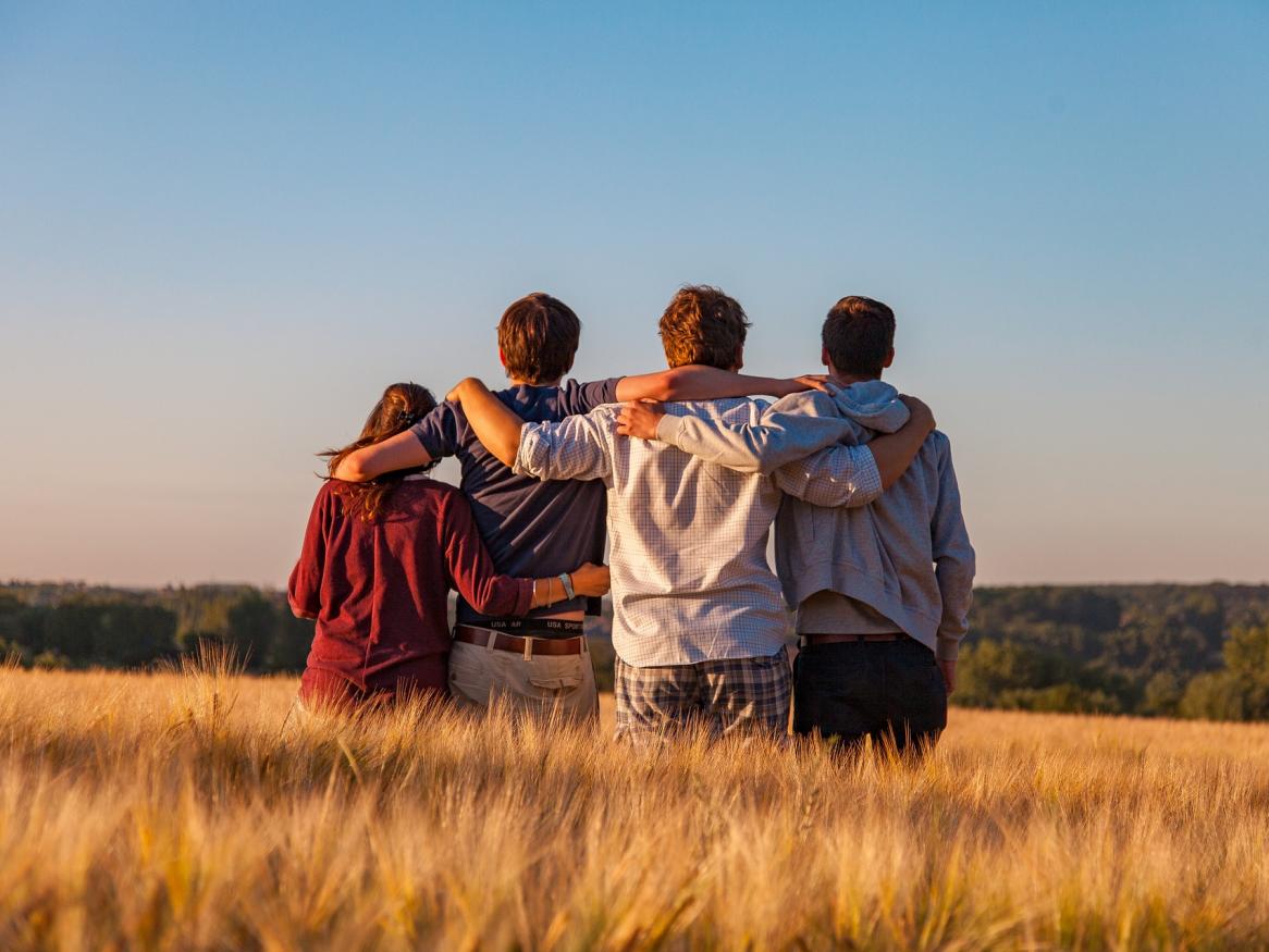 A young woman and three men standing in a field facing the sunset, arms around eachother