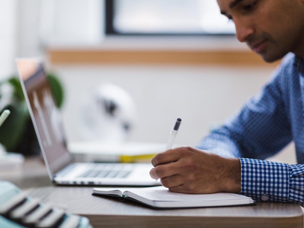 A person sitting at a desk with a laptop and writing in a notebook.