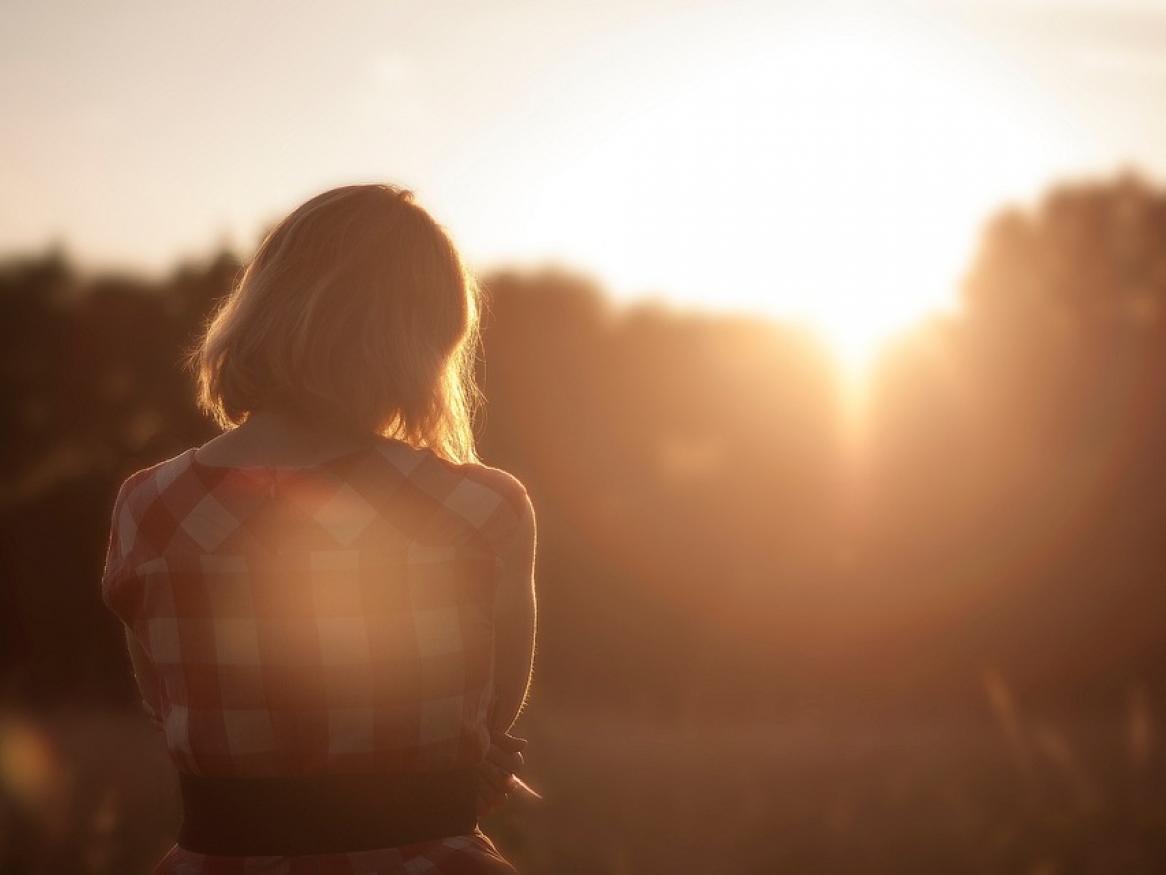 The back of a woman looking at the sunset over forest trees.