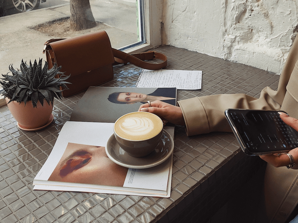 A cup of coffee, magazine, bag, and plant pot placed neatly on a cafe table